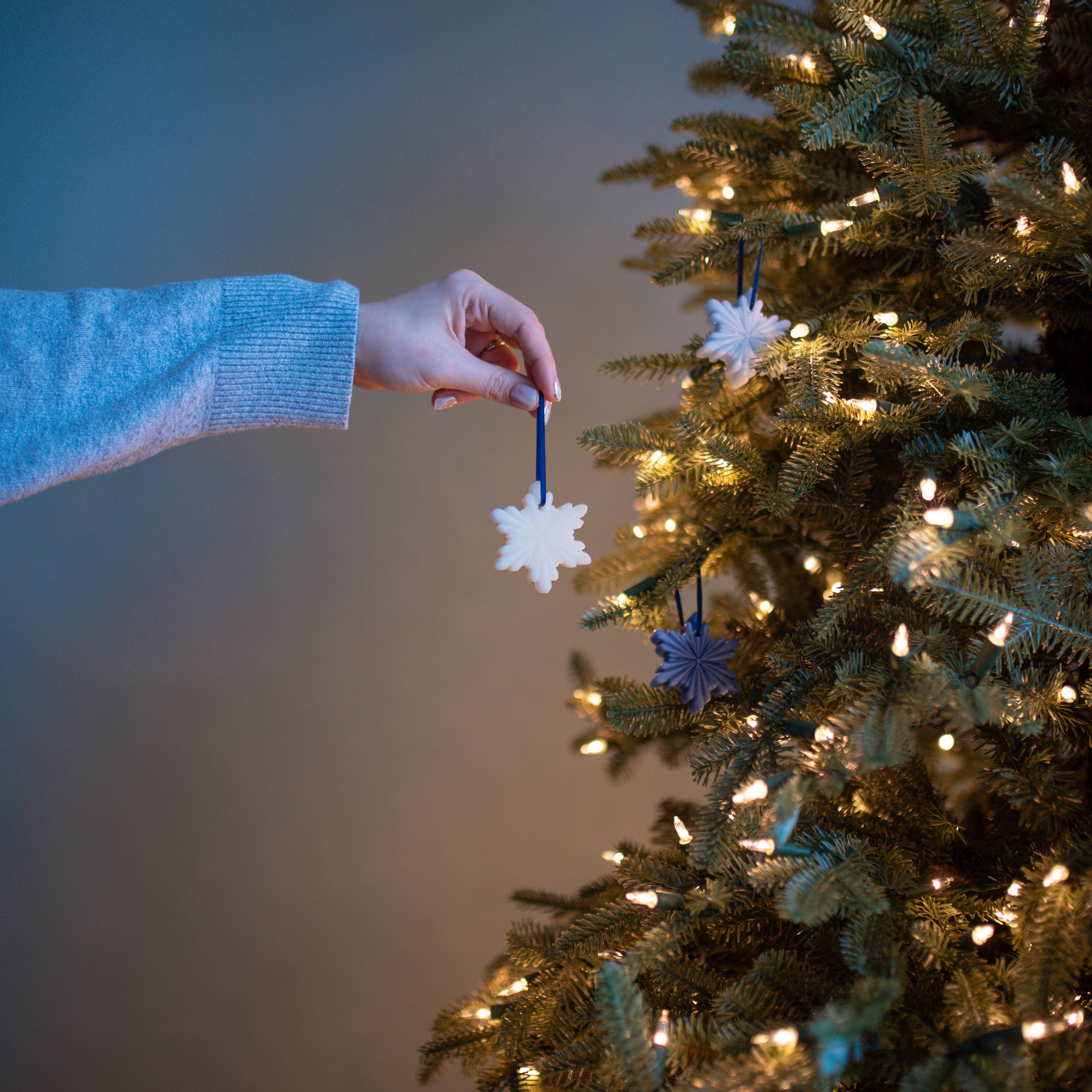 A hand placing a snowflake beeswax ornament on a lit christmas tree.